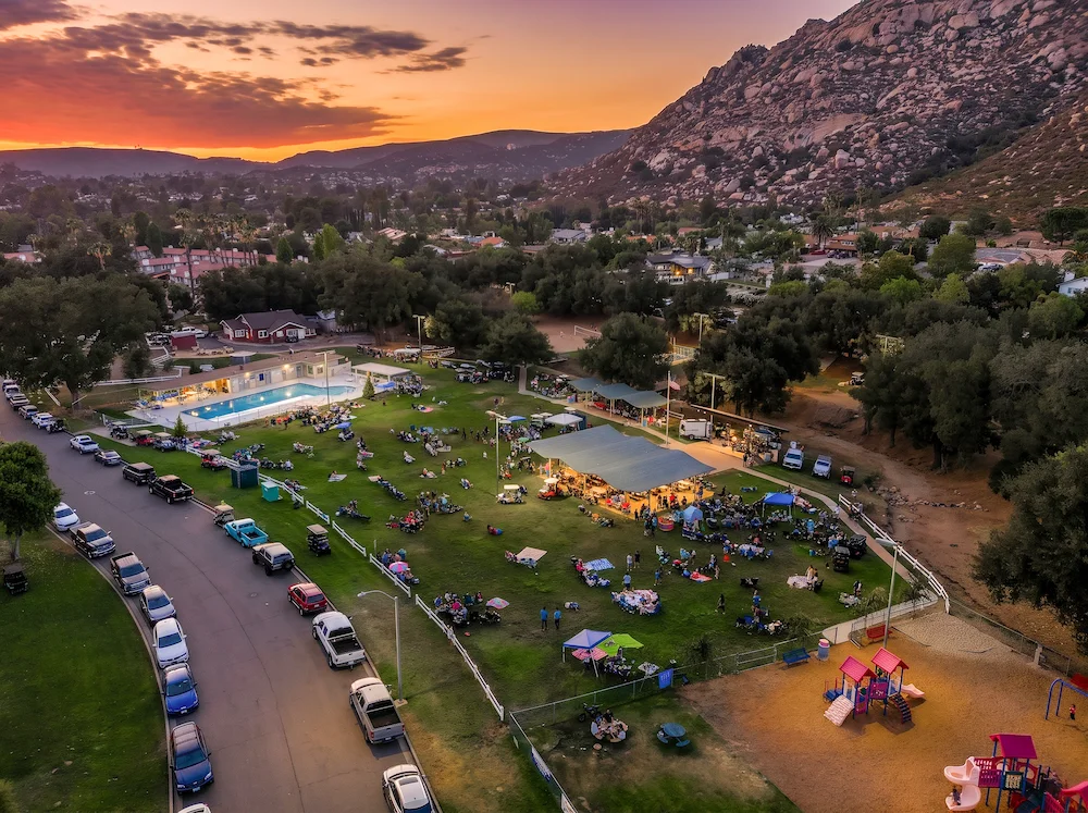Aerial view at sunset of a summer concert in the park at San Diego Country Estates, with neighbors gathered on the green, the pool, and the granite mountains behind.